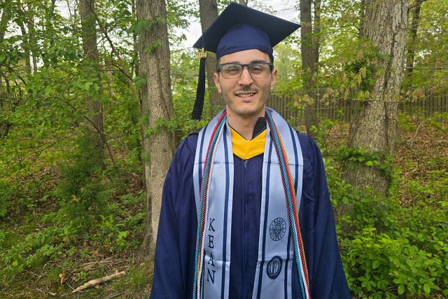 Kevin Hannon, in his blue 爱游戏体育 graduation cap and gown, stands in front woods.