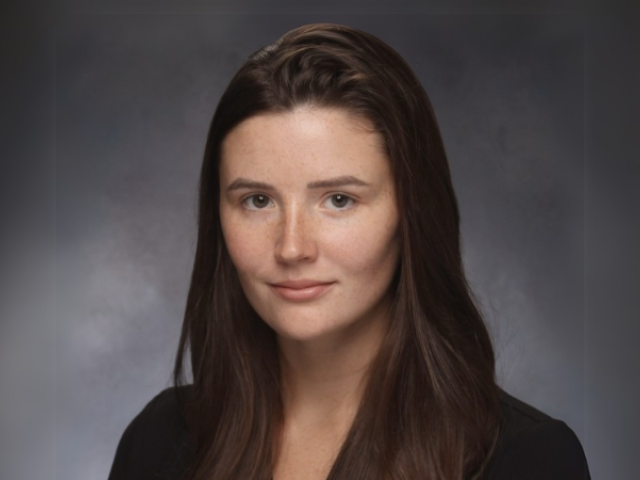 Headshot of white female with brown hair, back top; gray background
