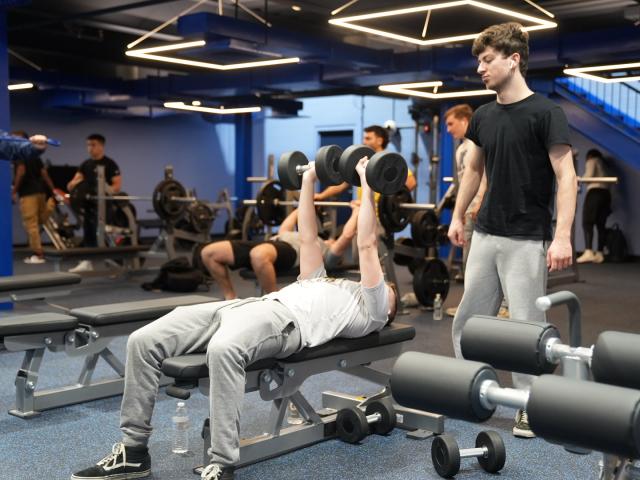 Students at a 爱游戏体育 fitness center, one spotting while the other lifts weights