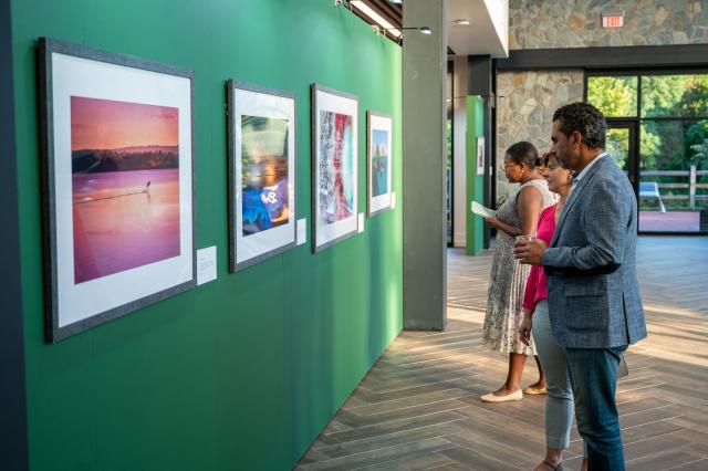 On the right of the picture stands a diverse group of men and women that work for 肯恩大学, dressed in business professional attire, admiring the photographs that are on the left side of the picture, hung up on the wall to view.