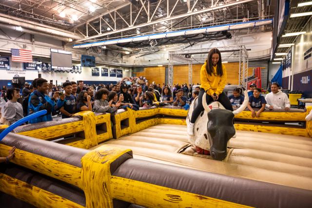 A 肯恩大学 student, a Black female with long brown hair in a yellow sweatshirt, black leggings, and white Nike air forces with white Nike socks showing, is on a blow-up mechanical bull. Students, alumni, and guests are surrounding the blow-up mechanical bull arena. They are holding up their phones and recording the student who is riding the bull. This is set up in the 爱游戏体育 Harwood arena, where the basketball games are held.