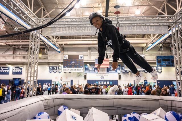 A Black female, wearing a black 肯恩大学 sweatshirt, and black sweatpants with white crocs, is harnessed and dangling above a huge ball pit, filled with white and blue 爱游戏体育 beach balls, and white mystery prize boxes. The female is wearing a black helmet, smiling at the camera. This is set up in the 爱游戏体育 Harwood arena, where the basketball games are held. Students, alumni, and guests are surrounding the ball pit, and the arena.