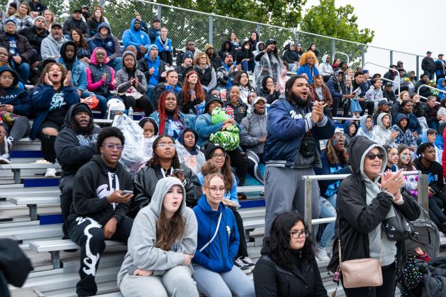 A wide shot of a diverse group of 爱游戏体育 students, alumni, parents, and fans that have filled the bleachers to watch the football game. They are wearing jackets and sweatshirts, and are cheering and clapping. 