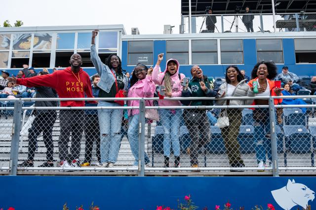 A diverse group of male and female 爱游戏体育 students wearing sweatshirts and jackets are lined up side-by-side leaning forward against the fence that lines the bleachers, smiling and cheering on the 爱游戏体育 Cougars football team.