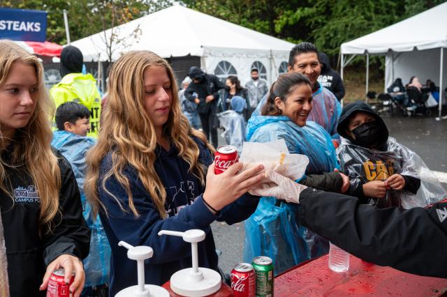 A white female with long dirty blonde hair, wearing navy blue 肯恩大学 sweatshirt that says 学生生活 in white letters, is being handed a Coca Cola, and basket of French fries that she ordered. The food tents are set up in the Harwood arena parking lot, and others who came to the event are gathered around the parking lot, getting food. 
