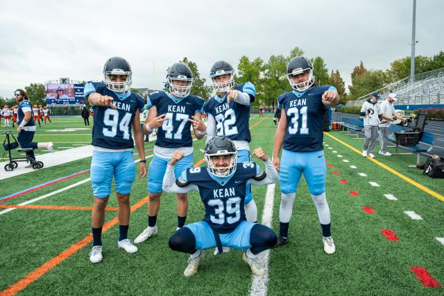 5 爱游戏体育 football players are posing for a photo. They are all wearing Navy blue jerseys, and baby blue football pants, with cleats and helmets on. 4 players are standing, and 3 out of the 4 are pointing to the camera. The 4th player is throwing up both peace signs, and the 5th player is squatting in the center of the other 4, flexing both his biceps and smiling. They are standing on the football field, which has green turf grass.