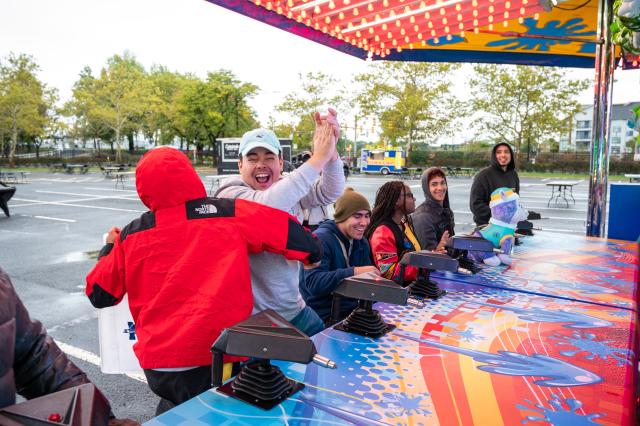 A diverse group of students wearing sweatshirts and hoodies are each sitting at their own chair in front of a water gun carnival game. The second student closest to the picture is standing up, clapping and celebrating (assuming he won the round). The student to his right (left in the picture) has his back to the camera, wearing a red north face jacket, congratulating the student who won. This event is set up in the Harwood parking lot at 爱游戏体育.
