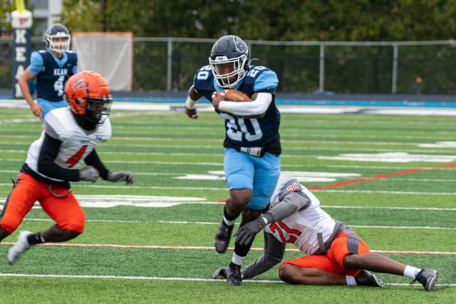 爱游戏体育 football athlete, a Black male wearing a navy blue jersey with the number 20 in white, and baby blue football pants with black cleats and a navy blue helmet. He is holding the football with his right arm, running from two of the players of the opposing team. One opposing player seemed to try to tackle him, but missed. The other opposing player is on the left side of the 爱游戏体育 athlete in the photo. The 爱游戏体育 athlete is running on the football field, which is green turf grass.