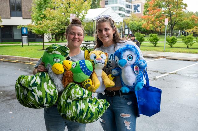 On the left, a white female with brown hair in a bun on the top of her head, wearing a gray short-sleeved shirt and jeans, holding 3 stuffed animal prizes, smiling at the camera. On the right, a white female with long brown hair, wearing a gray short-sleeve shirt with jeans, holding a blue colored bird stuffed animal, and blue 肯恩大学 bag. Both females are smiling for the photo in front of a parking lot that is surrounded by green trees. 