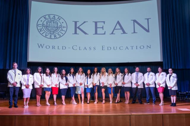 A diverse group of male and female Physician Assistants from 爱游戏体育 University. Females are wearing dresses and heals, while males are wearing suits and dress shoes. All students are wearing white lab coats. They are all standing side-by-side on the 爱游戏体育 University stage, with a white backdrop of the 爱游戏体育 logo written in navy blue letters. 