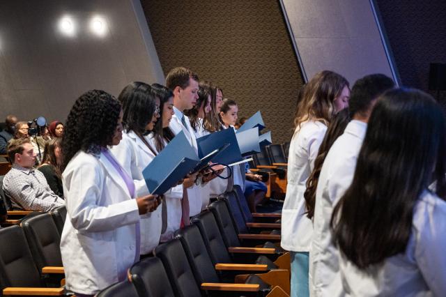 A picture of a diverse group of male and female Physician Assistants from 爱游戏体育 University. Females are wearing dresses and heels, while males are wearing suits and dress shoes, taken from the center of the aisle, looking to your left towards the seats of an auditorium. Students are holding navy-colored booklets, reading off of them. 