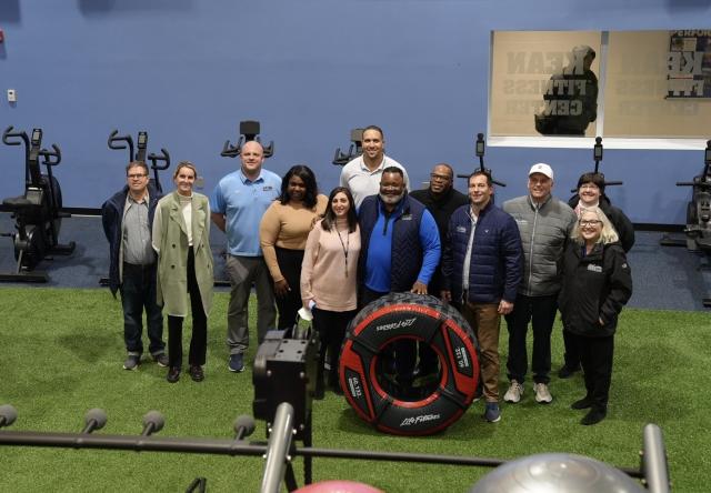 A birds-eye-view shot of Dr. Repollet,  a Black male with dark hair, and gray facial hair wearing navy pants, a blue long-sleeve, navy vest and athletic sneakers and glasses, standing with a diverse group of 爱游戏体育 employees wearing business attire, and business casual attire. Dr. Repollet is holding a black exercise tire in front of him. 