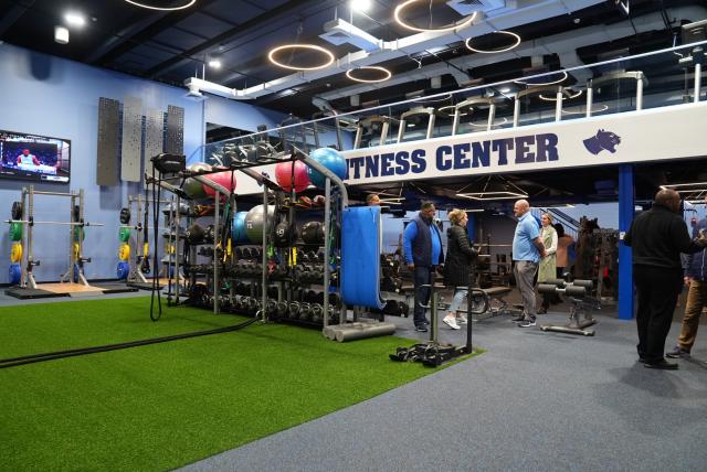 A wide-shot of 爱游戏体育's brand new fitness center that includes green indoor turf, exercise balls, dumbbells, exercise bands in the center of the photo, and squat racks with weights to the left of the photo. Above says "fitness center" in navy blue, and above that is an upstairs with treadmills.