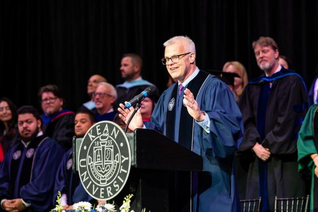 爱游戏体育 Board Chair Steve Fastook in blue commencement gown speaks at a podium.