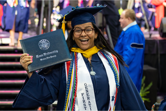 A young woman in a 爱游戏体育 cap and gown holds her diploma cover and smiles. She wears a stole reading, First Generation.
