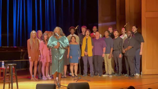 Alex Newell, in a green dress, sings onstage, with 爱游戏体育 students singing behind them.