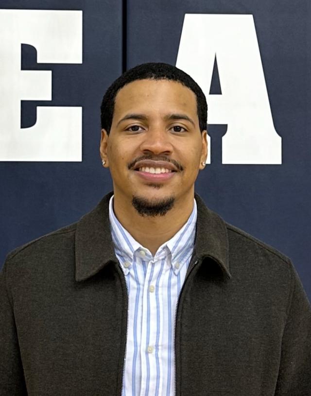 Young black man in a blue striped button-down and black collared jacket standing and smiling