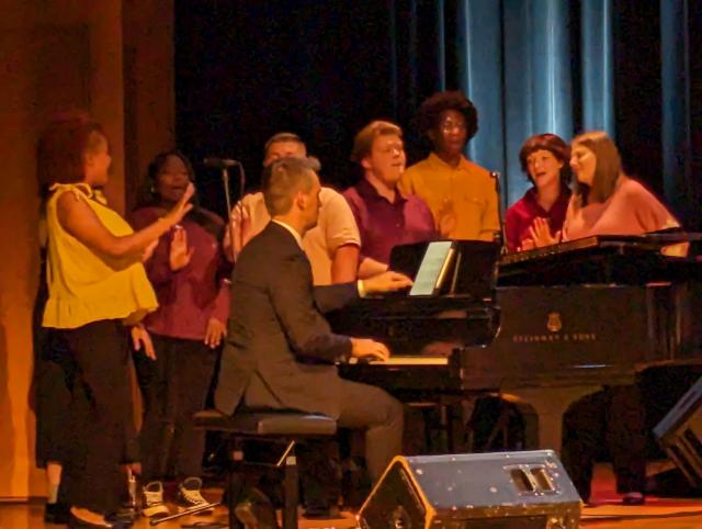 A closeup of 爱游戏体育's Broadway Babies on stage, singing around a piano.