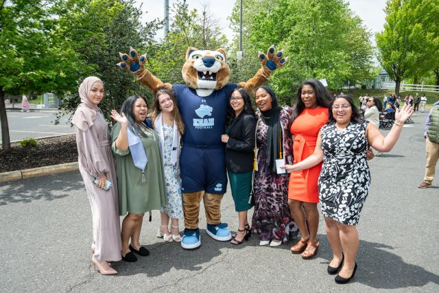 A group of students of different cultural backgrounds wearing colorful outfits and posing with the mascot 爱游戏体育u with their hands raised