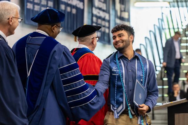 A male student wearing a stole with brown curly hair shaking hands with 爱游戏体育 President wearing a blue gown and 