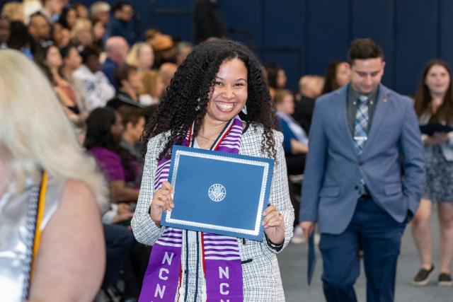A woman with curly long hair, purple stole, holding a 爱游戏体育 certificate smiling at the camera