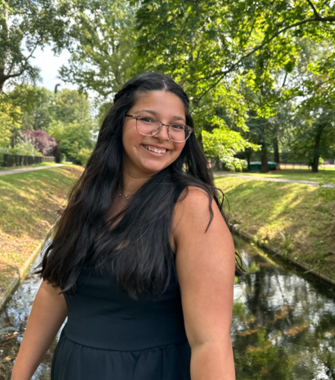 Photo of Esther Gonzales in front of a river and surrounded by trees. Esther is wearing glasses and a black dress.