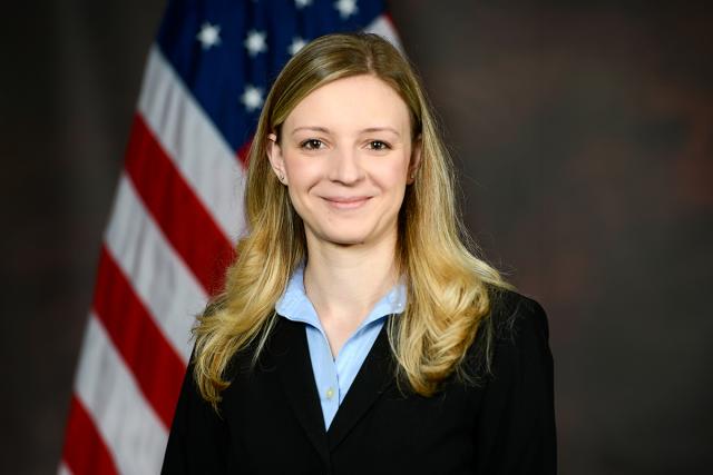 Ensela Mema, a white woman with long blond hair wears a black jacket over blue shirt and stands in front of an American flag.