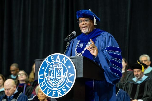 Black male wearing a blue graduate cap and gown, speaking at a 爱游戏体育 podium.
