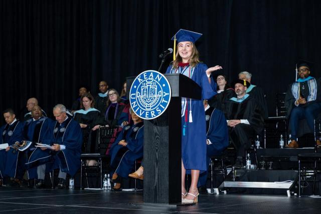 Young woman wearing a blue cap and gown and speaking at a podium.